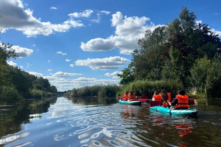 Auf dem Foto sind zwei Kajak-Boote zu sehen mit je drei Insassen. Sie paddeln auf einem Fluss zwischen Bäumen entlang bei blauem Himmel.