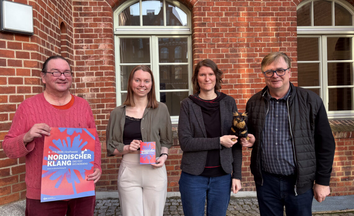 Das Team des Nordischen Klangs steht für ein Gruppenbild nebeneinander und hält Flyer, Plakat und Plüschdackel in den Händen.