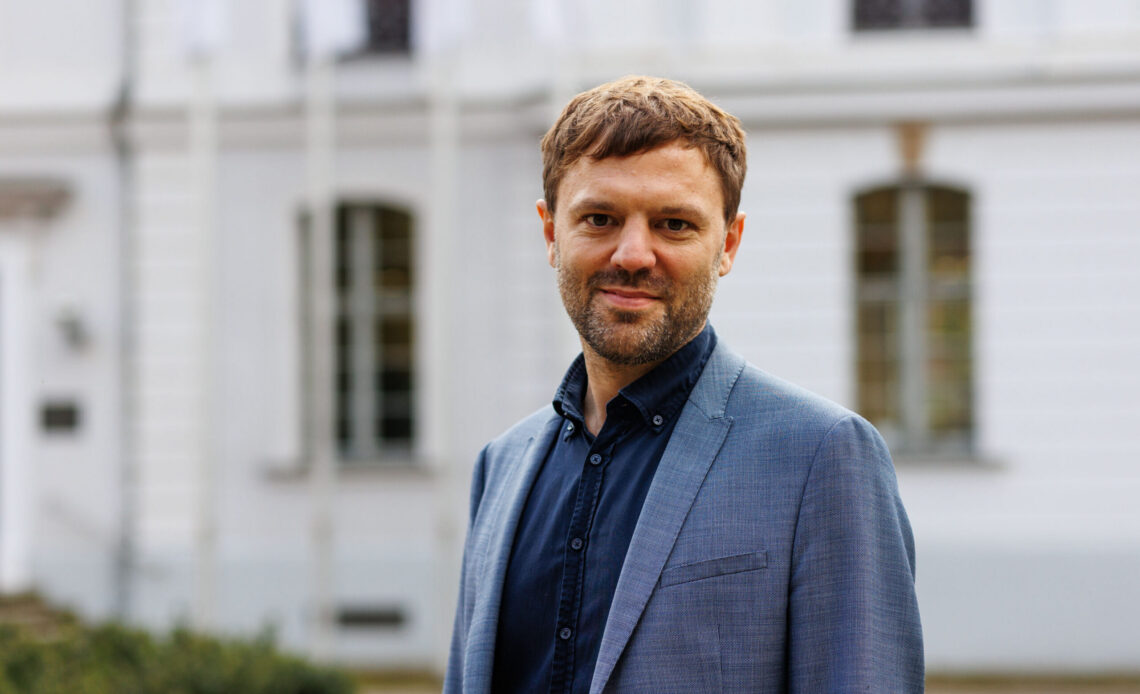 Das Portrait zeigt Prof. Dr. Jan-Hinnerk Antons. Er hat braune kurze Haare, trägt einen blauen Anzug und lächelt freundlich in die Kamera. Im Hintergrund ist ein weißes Gebäude zu sehen.