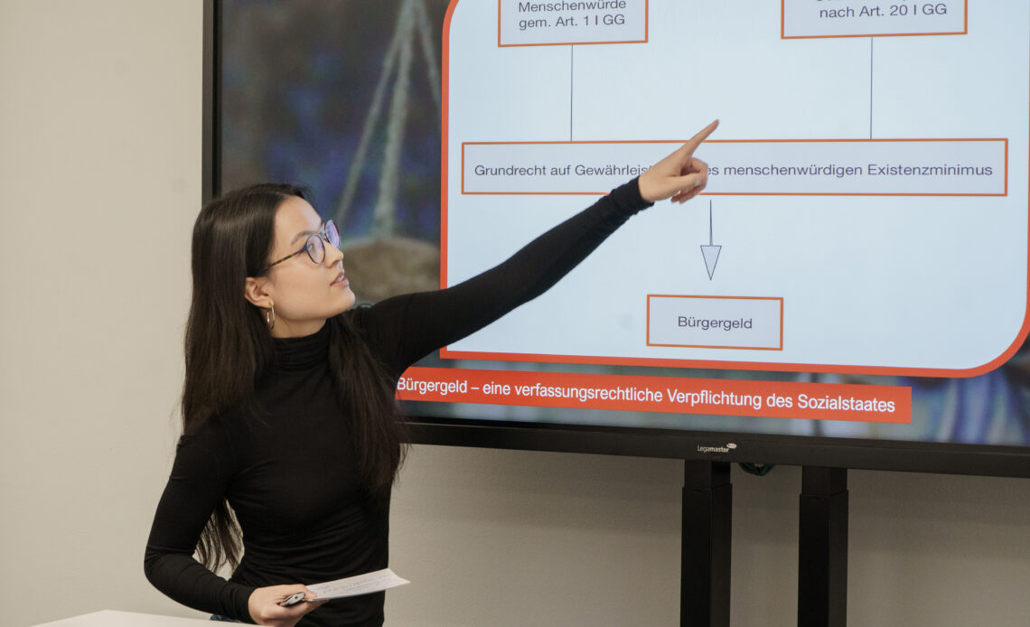 Eine junge Frau in Schwarz deutet auf eine Infografik, die Informationen zum deutschen Sozialstaatsprinzip und Bürgergeld enthält.