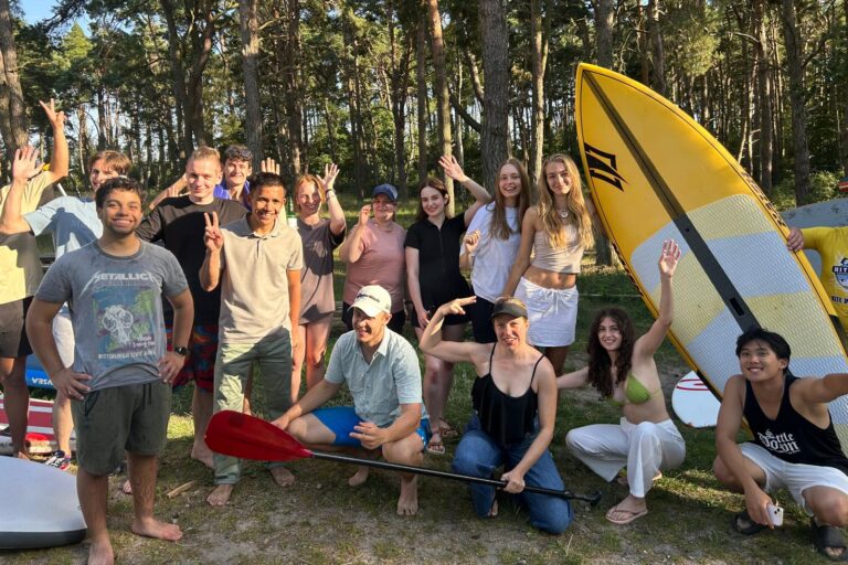 Das Foto zeigt die Gruppe der Teilnehmenden, wie sie vor einem Wald zusammenstehen und begeistert in die Kamera schauen. Zwei von ihnen halten ein Paddel und auch ein Stand-Up Paddle Board ist zu sehen.