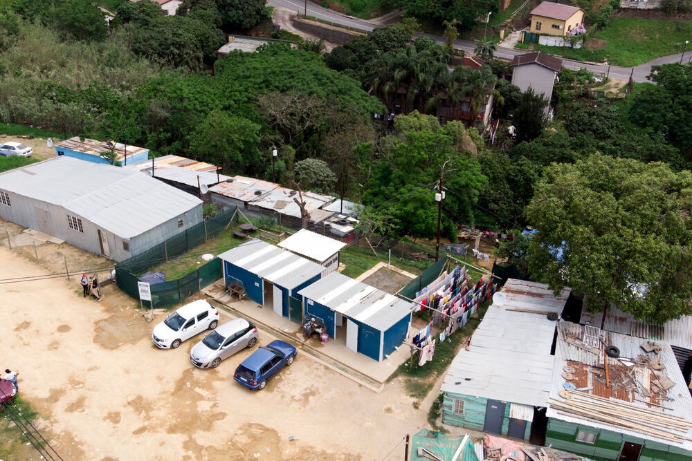 Das Foto ist aus der Vogelperspektive aufgenommen und zeigt einige Häuser mit Wellblechdächern. Hinter den Häusern stehen einige Bäume und am oberen Bildrand ist eine Straße zu sehen.