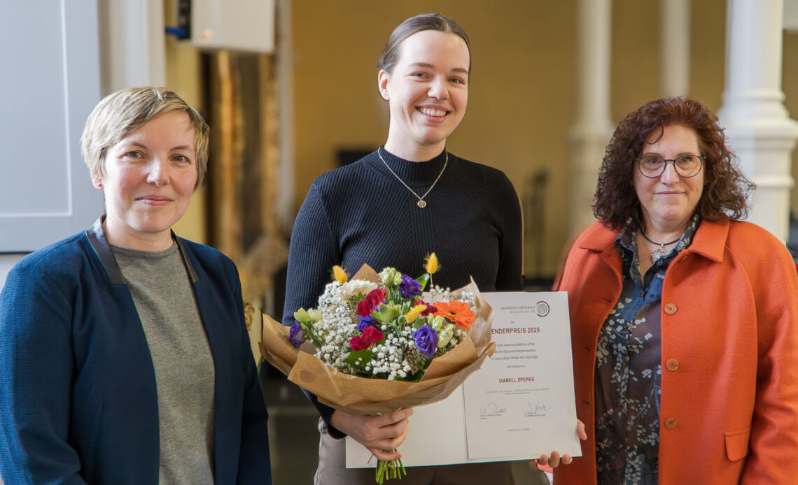 Das Foto zeigt drei Frauen, die freundlich in die Kamera lächeln. Die Frau in der Mitte ist jünger als die anderen beiden und hält einen Blumenstrauß und eine Uhrkunde in den Händen.