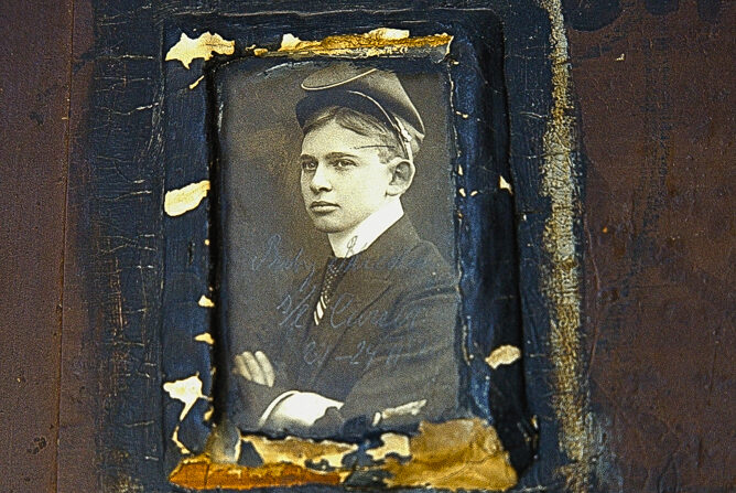 Das Foto zeigt ein sehr altes schwarz-weiß Foto von einem jungen Mann (Bubi). Der Mann ist im Seitenprofil zu sehen, trägt eine Kopfbedeckung und einen dunklen Anzug.