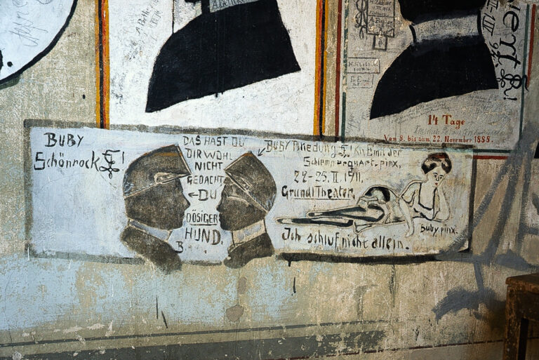 Das Foto zeigt eine Zeichnung an der Wand des Karzers. Es sind Seitenprofile zweier Männer zu sehen, die sich gegenüberstehen. Zusätzlich wird eine Art Gespräch zwischen den beiden abgebildet. Neben den beiden Gesichtern ist außerdem eine leicht bekleidete, liegende Frau abgebildet, die Inhalt des Gesprächs zwischen den beiden Männern darzustellen scheint.