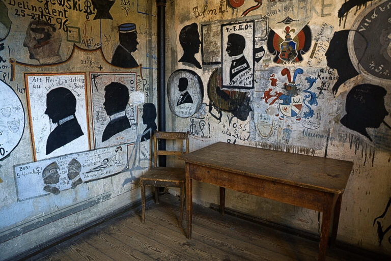Das Foto zeigt einen Raum, an dem die Wände mit zahlreichen Zeichnungen übersäht sind. An einer der Wände steht ein alter Holztisch und ein Stuhl. An den Wänden stechen vor allem schwarze Zeichnungen hervor, die das vereinfachte Seitenprofil von Männern zeigen.