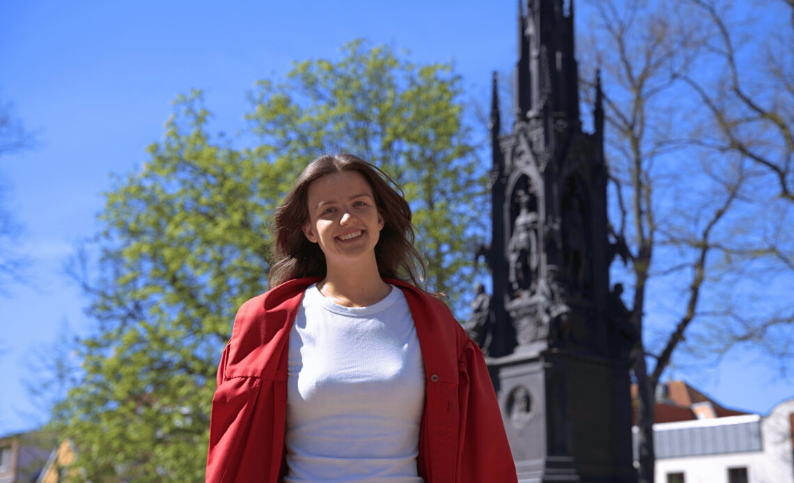 Das Foto zeigt eine junge, braunhaarige Frau, die mit einem bereiten Lächeln in die Kamera schaut. Sie trägt ein weißes Oberteil und darüber eine rote Jacke. Im Hintergrund ist ein historisches Denkmal zu sehen, mehrere Bäume und klarer, blauer Himmel.