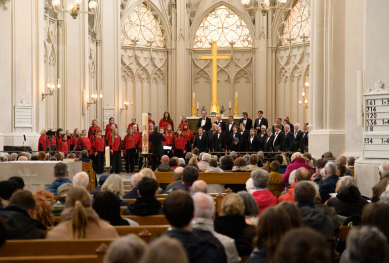 Das Foto zeigt den Greifswalder Dom von innen. Vorne am Altar steht ein Chor vor einem großen goldenen Kreuz. Der Chor ist farblich in zwei Seiten aufgetrennt. Die eine Hälfte trägt rote Oberteile und schwarze Hosen, die andere Hälfte ist ganz in schwarz gekleidet.