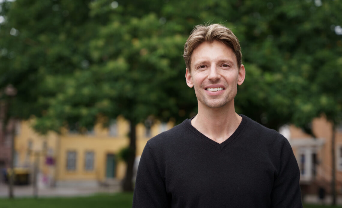 Das Foto zeigt einen braunhaarigen, lächelnden Mann mit kurzen Haaren. Er trägt ein schwarzes Oberteil und im Hintergrund sind Häuser und Bäume zu sehen.
