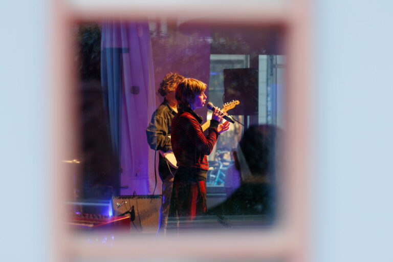Das Foto zeigt eine Sängerin im Seitenprofil, wie sie gerade in ein Mikrofon singt. Schräg hinter ihr ist eine Person zu sehen, die die Sängerin an einer Gitarre begleitet.