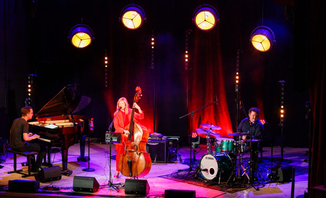 Das Foto zeigt eine Bühne auf der drei Musiker:innen stehen. In der Mitte steht eine Frau am Cello, rechts von ihr ein Mann am Schlagzeug und links von ihr spielt ein Mann Klavier.