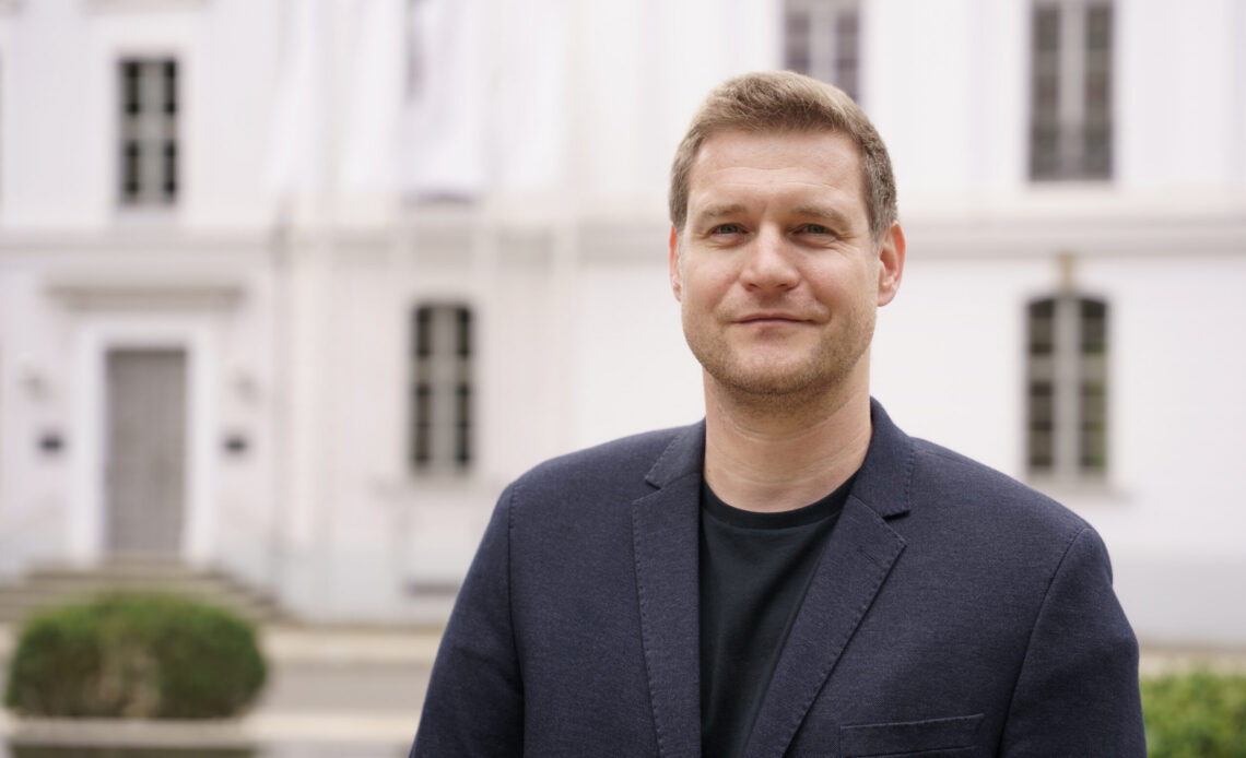Das Foto zeigt einen Mann mit kurzen braunen Haaren, der freundlich in die Kamera lächelt. Er trägt ein blaues Jackett und im Hintergrund ist ein weißes, historisches Gebäude zu sehen.