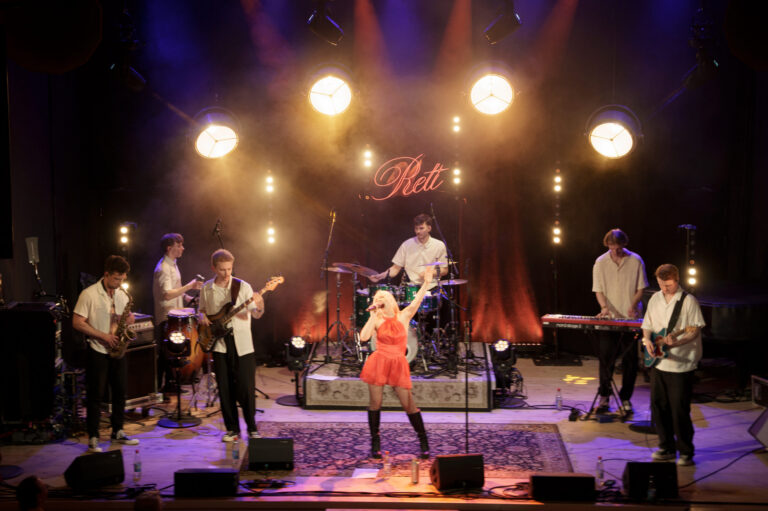 Das Foto zeigt eine 7-köpfige Band auf einer Bühne. In der Mitte steht eine junge Sängerin in einem rotem Kleid, im Halbkreis hinter ihr stehen sechs Männer in weißen Oberteilen, die Instrumente spielen.