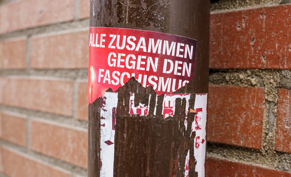 Das Bild zeigt eine Laterne, die mit mehreren zerrissenen Stickern beklebt ist. AUf einem steht: "Alle zusammen gegen den Faschismus". Die anderen Sticker sind so abgekratzt, dass kein text zu erkennen ist.