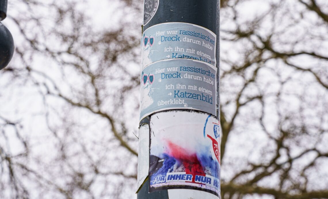 Das Bild zeigt eine Laterne auf der mehrere Sticker kleben. Im Fokus stehen zwei hellblaue Sticker auf denen steht: "Hier war rassistischer Dreck, darum habe ich ihn mit einem Katzenbild überklebt". Zudem ist eine weiße Kattze mit Sonnenbrille abgebildet. Drunter klebt ein blau, weiß, roter Hansa Rostock Aufkleber.