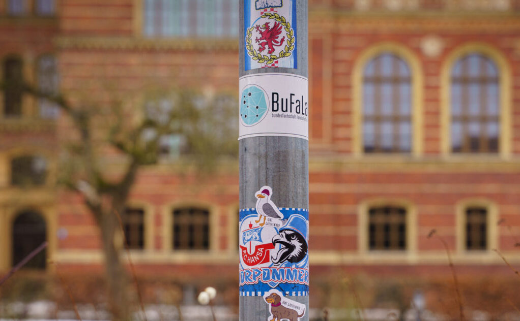 Mehrere Sticker kleben auf einem Laternenmast. Im Hintergrund ist ein Backsteingebäude.