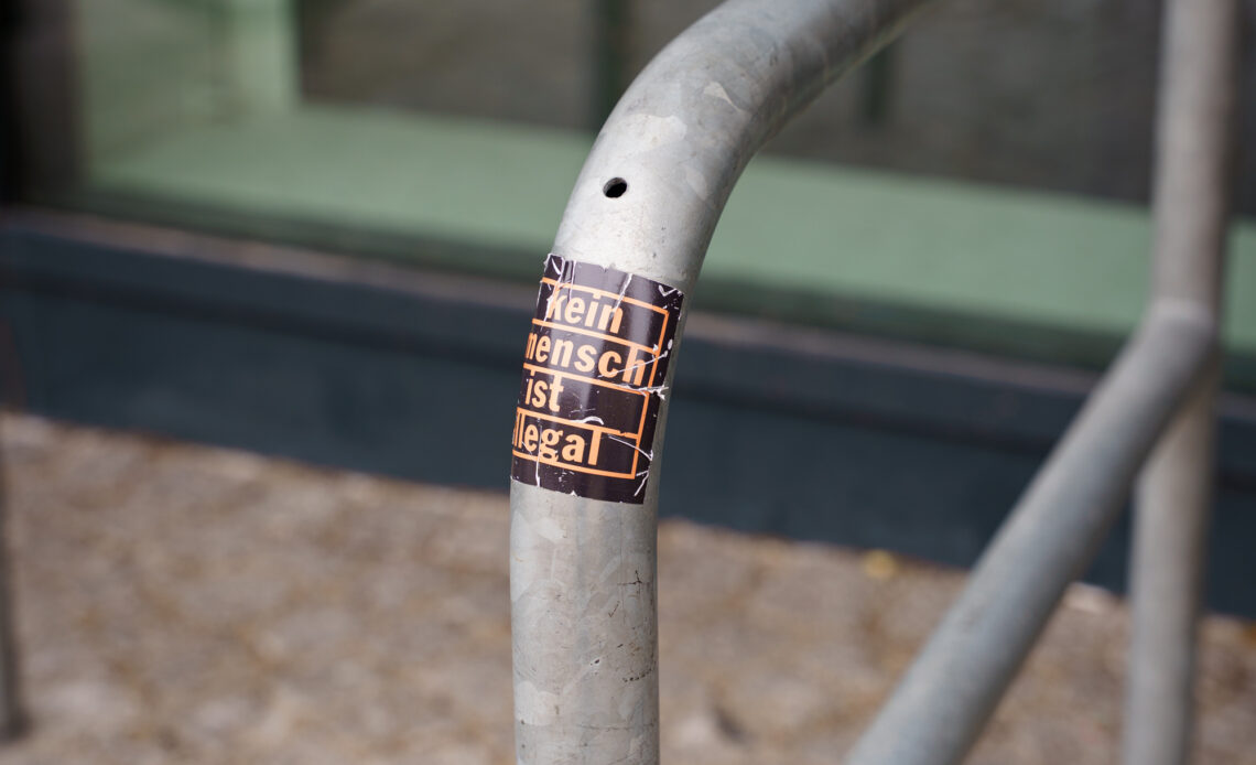 Ein Sticker mit dem Spruch "Kein Mensch ist illegal" klebt auf einem Fahrradständer. Im Hintergrund ist eine spiegelnde Glasscheibe