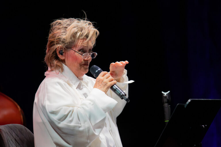Das Foto zeigt die Sängerin Gitte Hænning im Seitenprofil, während sie in ein Mikrofon sinkt. Sie hat kurze, helle Haare, trägt eine Brille und eine weiße Bluse.
