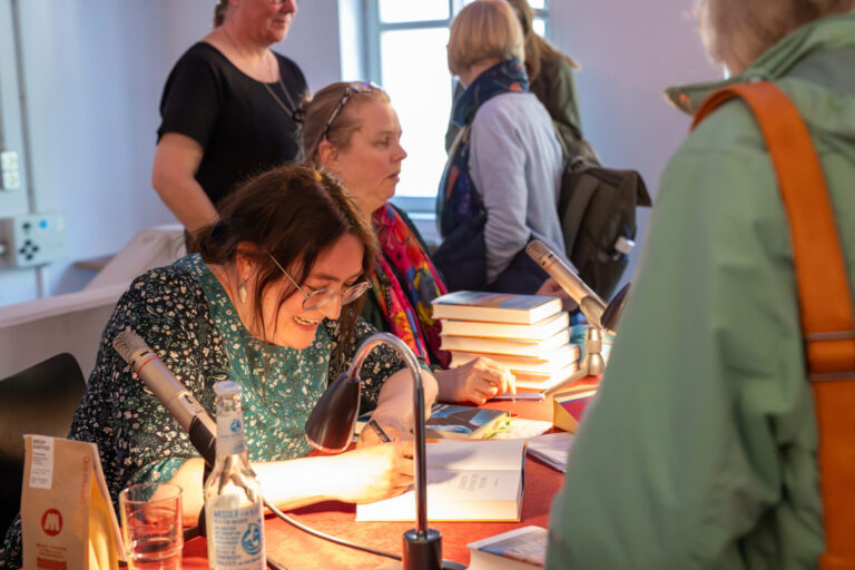 Das Foto zeigt einen Raum, in dem sich mehrere Menschen befinden. Im Vordergrund sitzt eine Frau an einem Tisch und signiert gerade ein Buch. Neben ihr steht ein kleiner Bücherstapel.
