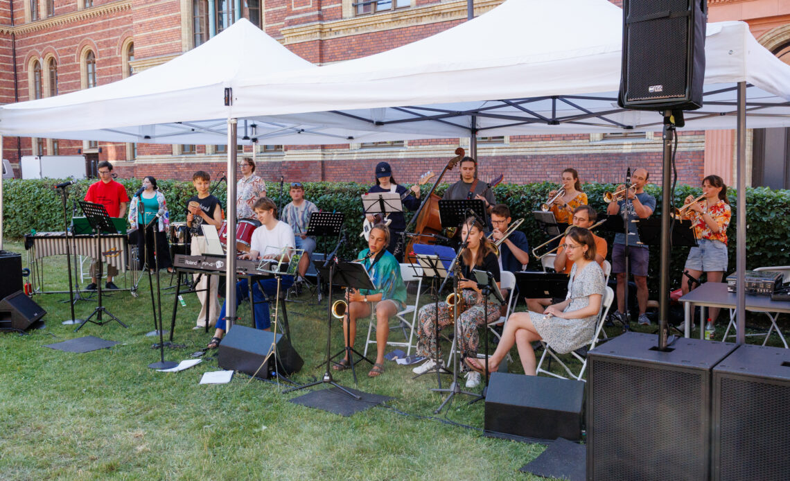 Die UniBigBand steht unter einem Zelt und spielt auf verschiedenen Instrumenten Musik.