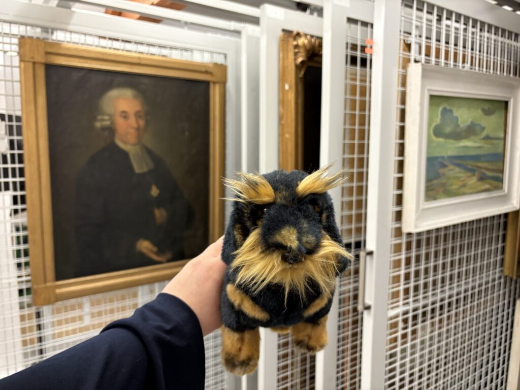 Ein schwarz-brauner Kuscheltier-Dackel wird von einer Hand vor ein Gemälde gehalten. Das Gemälde zeigt einen Mann. Am rechten Bildrand sind weitere Gemälde mit verschiedenen Motiven zu erkennen.