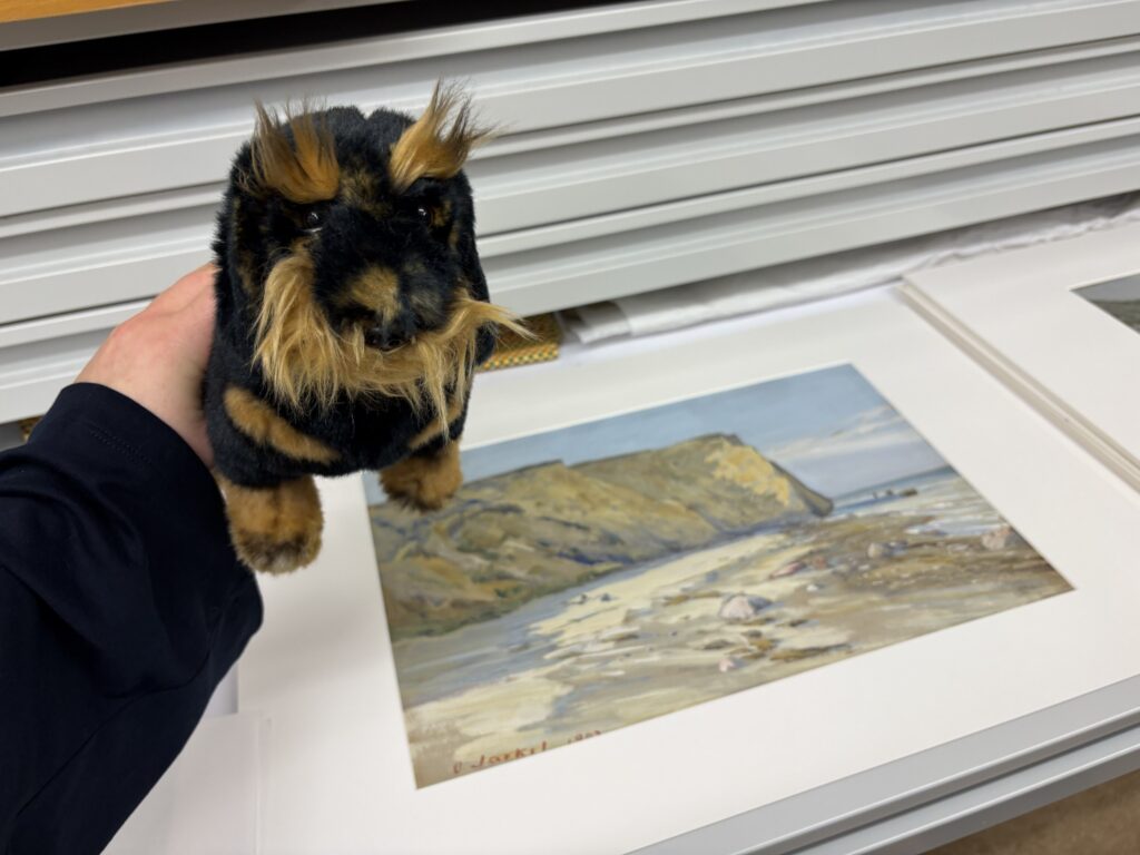 Ein schwarz-brauner Kuscheltier-Dackel wird von einer Hand über ein Gemälde gehalten. Das Gemälde zeigt eine Strandlandschaft.