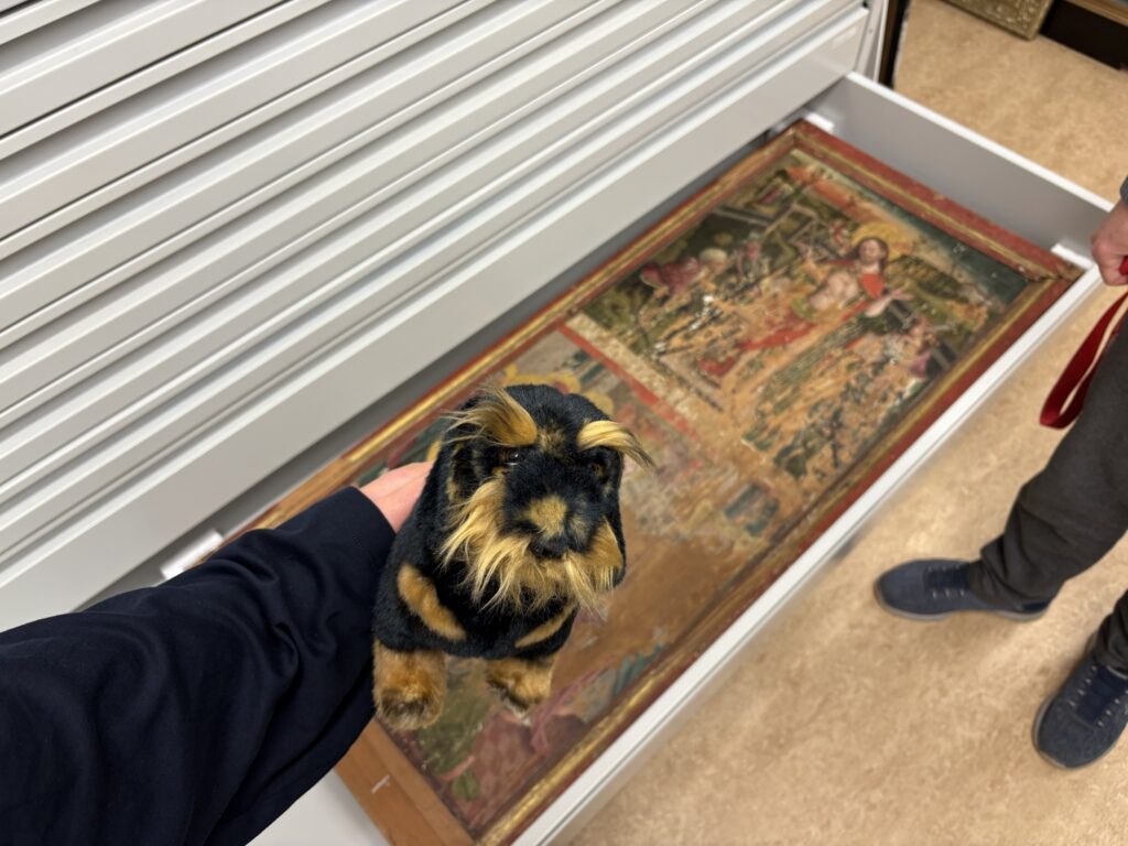 Ein schwarz-brauner Kuscheltier-Dackel wird von einer Hand über eine geöffnete Schublade gehalten. In der Schublade befindet sich ein altes, abgenutztes Gemälde auf einer Holztafel.