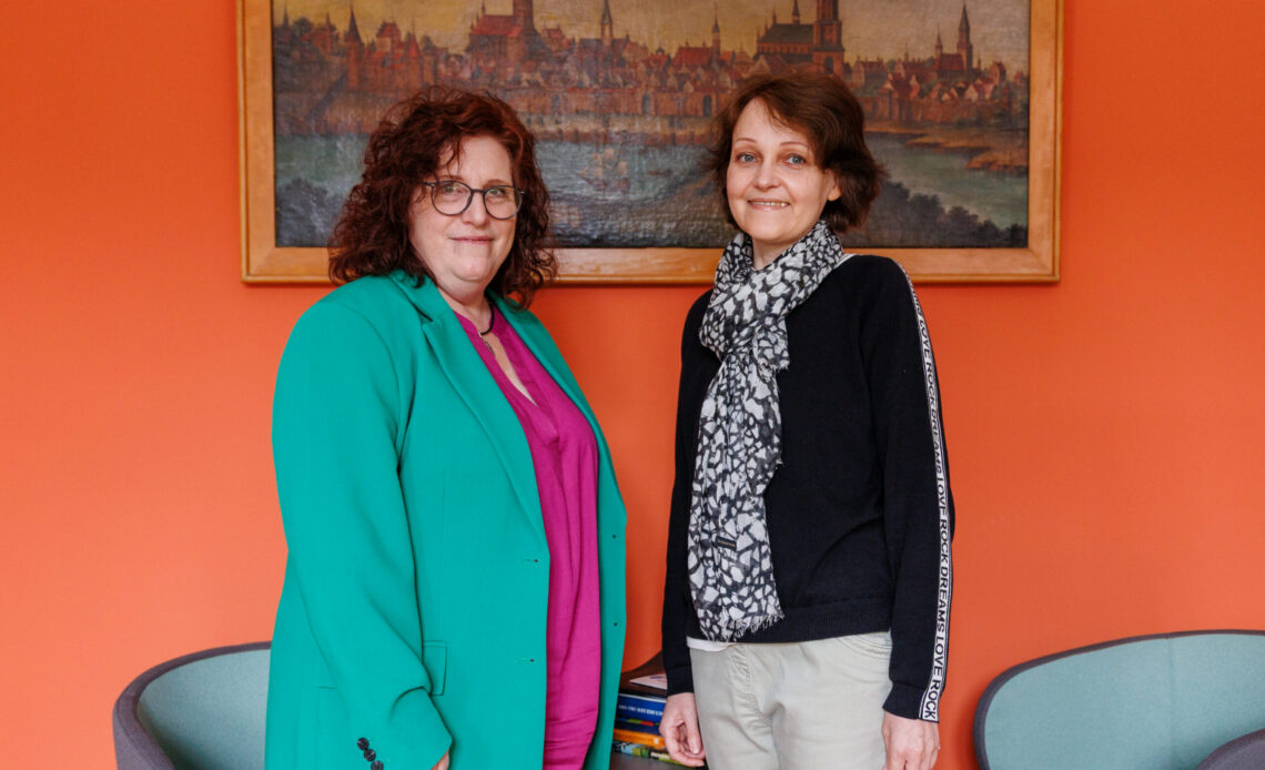 Das Foto zeigt zwei Frauen mittleren Alters, die nebeneinander stehen und in Kamera lächeln. Die Frau rechts hat braune kurze Haare, und trägt ein schwarzes Oberteil mit einem schwarz, weißem Schal. Die Frau links hat braune lockige, lange Haare, trägt eine Brille und ein türkises Jackett.