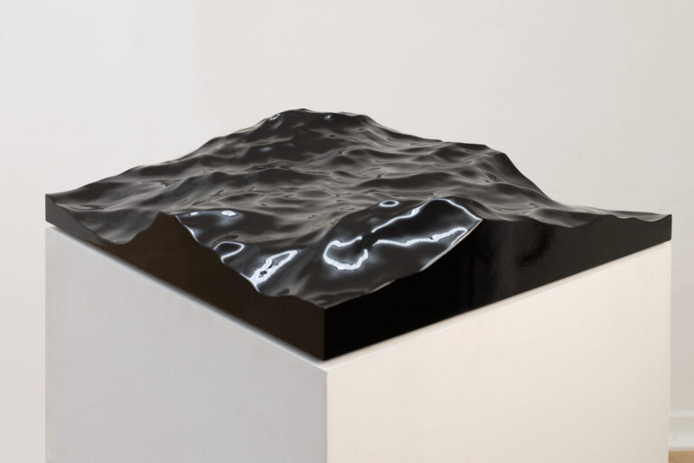 Das Foto zeigt ein Skulptur. Die Skulptur besteht aus einem weißen Kasten auf dem eine schwarze wellige Oberfläche angebracht ist.