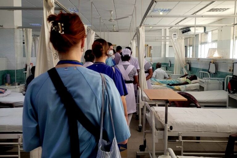 Das Foto zeigt einen Zimmer, in dem auf der rechten und linken Seite Krankenbetten stehen. Die Betten werden durch einen Gang getrennt, durch den gerade medizinisches Personal läuft.