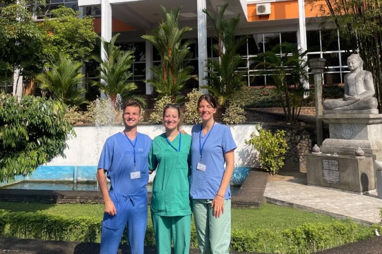 Das Foto zeigt drei Menschen in Krankenhauskleidung, die vor einem Gebäude und kleinen Palmen in die Kamera lächeln.