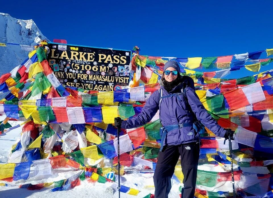Das Foto zeigt eine Frau in dicken Wintersachen, wie sie breit lächelnd vor einem bunten Schild in den Bergen steht. Auf dem Schild steht „Larke Pass“.