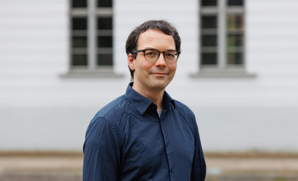 Das Portrait zeigt einen Mann mit dunklen, kurzen Haaren, der freundlich in die Kamera lächelt. Er trägt ein dunkelblaues Hemd und eine Brille. Im Hintergrund ist ein weißes Gebäude zu sehen.