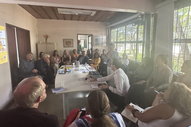 Das Foto zeigt einen Raum, in dem ungefähr 20 Menschen um einen Tisch sitzen und eine Besprechung abhalten.