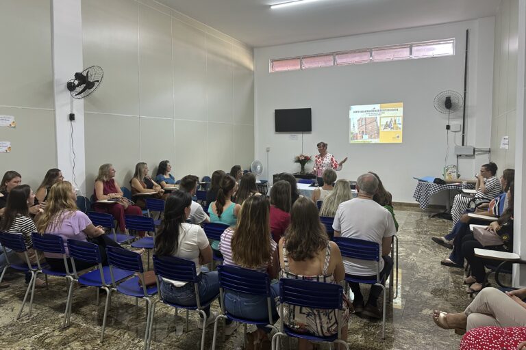 Das Foto zeigt einen Raum, in dem in mehreren Stuhlreihen Menschen sitzen und an die Tafel ganz vorne im Raum schauen. Dort steht eine Frau zwischen einem Bildschirm und einer PowerPoint und redet.